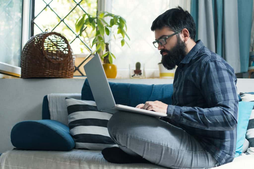 male developer wearing plaid shirt coding on a laptop from a couch