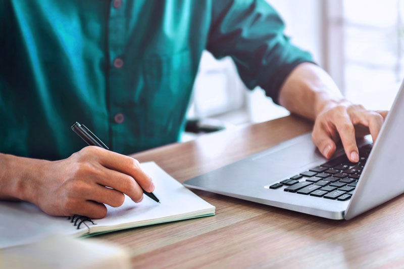person writing on notebook and using a laptop